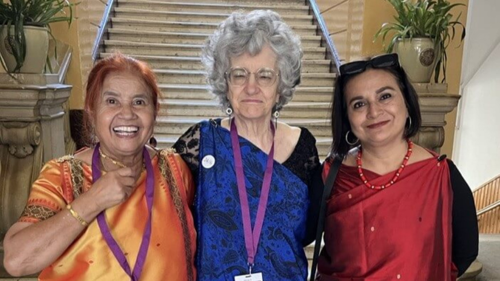 3 women: one with long red hair, dressed in bright orange, taller one in center with curly gray hair and wearing a purple dress, and the one on the right wearing shiny red, with medium-length dark hair. 2 wear event lanyards and nametags