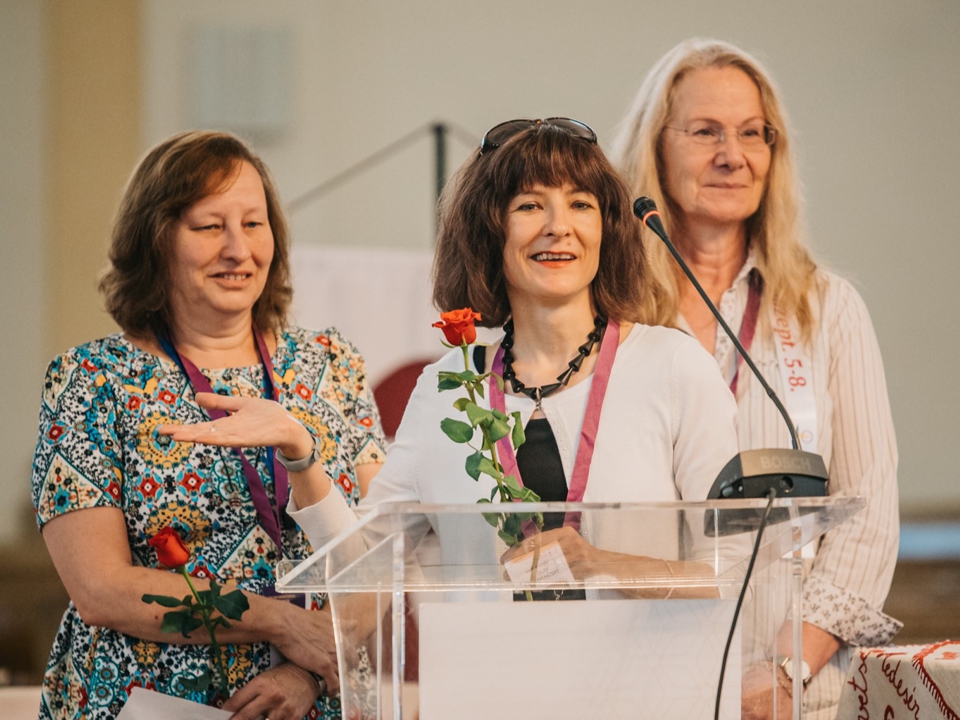 3 women: one with medium length red hair, dressed in a print blouse, a dark-haired woman in center holding a rose, and on the right a woman with long blonde hair wearing a long sleeve white blouse