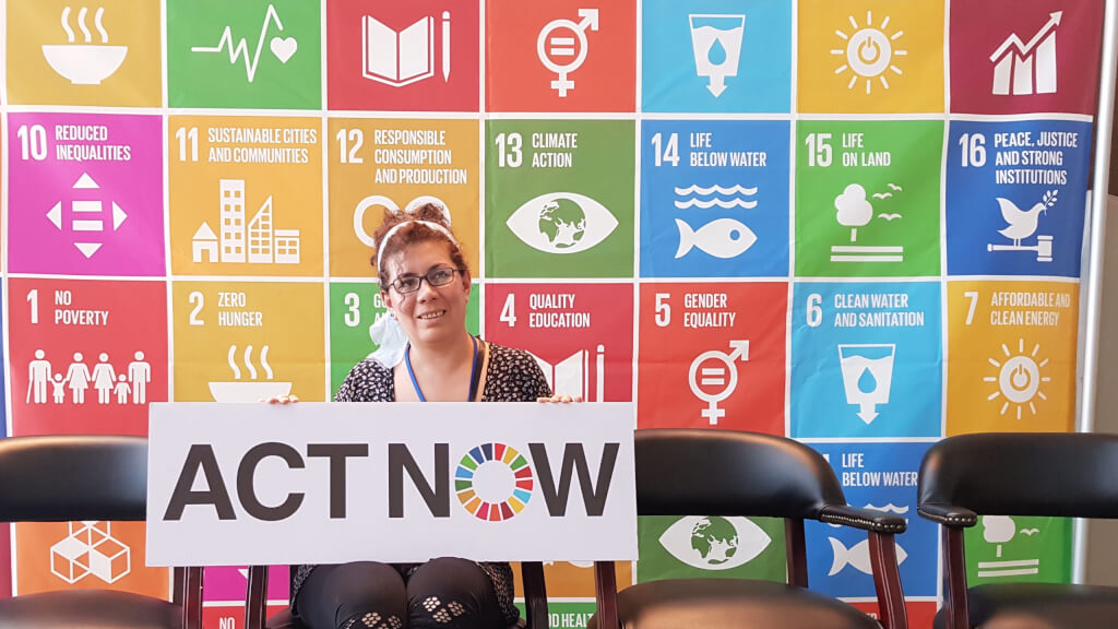 Carmen Capriles and the UN Sustainable Development Goals - a person holding an ACT NOW sign standing in front of a wall with a colorful grid of graphics